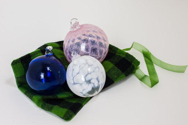 May include: Three glass ornaments in a green and black plaid bag with a green ribbon. The ornaments are blue, white with gray swirls, and pink with purple swirls.