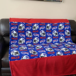 May include: A blue fleece blanket featuring a repeating pattern of Buffalo Bills football helmets and the word BILLS in red and white. The blanket has solid red fabric borders along the top and bottom edges, creating a distinct rectangular design.