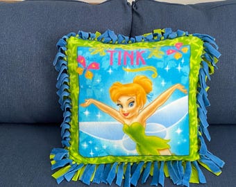 May include: A green and blue fleece pillow with a fringe border. The pillow features a colorful image of Tinkerbell with the word "TINK" printed on the pillow.