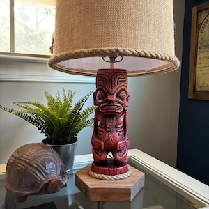 May include: A red tiki-style table lamp with a burlap shade and rope trim. The lamp features a carved wooden base and a decorative rope detail. A small fern and a wooden turtle are also visible.
