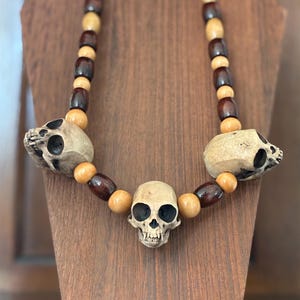 May include: A necklace featuring three realistic, off-white skull beads interspersed with alternating dark brown and light tan wooden barrel-shaped beads. The central skull is positioned facing forward, while the two side skulls face outward.