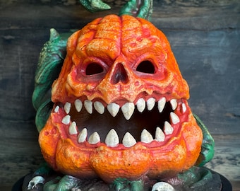 Könnte beinhalten: Eine orangefarbene Kürbisskulptur mit einem bedrohlichen Gesicht, scharfen Zähnen und einer grünen Ranke. Der Kürbis steht auf einem schwarzen Sockel mit dunkler, strukturierter Oberfläche, ideal für Halloween.