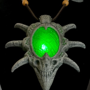 May include: A gray, fantasy-themed pendant necklace with a glowing green center. The pendant has a skull-like design with intricate details and is suspended from a brown cord with wooden beads and decorative axe-shaped accents.