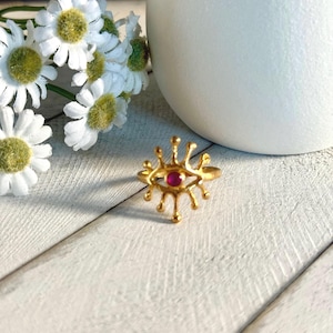 May include: A gold-colored ring with an eye-shaped design and a small, round, red gemstone in the center. The ring is set against a white background with white flowers and a white vase.