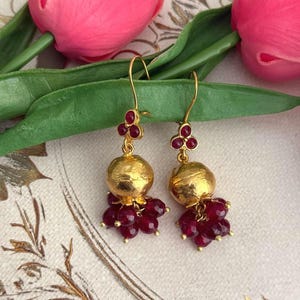 May include: A pair of gold-toned drop earrings featuring a small three-stone ruby cluster at the top, a central smooth gold-toned sphere, and a dangling cluster of faceted deep red ruby beads at the base, suspended from gold-toned ear wires.