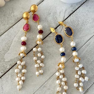 Peut inclure: Boucles d'oreilles dorées avec des pierres précieuses et des perles. Une paire présente des pierres roses et rouges, l'autre des pierres bleues et blanches. Les deux ont un groupe de petites perles suspendues en bas.