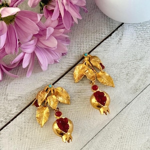 May include: Gold-toned earrings featuring a leaf and pomegranate design. The earrings have a turquoise accent and a red center in the pomegranate. The earrings are set against a white and gray wooden surface with pink flowers.