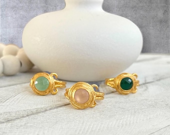 May include: Three gold-colored rings with different colored gemstones: green, pink, and dark green. The rings are in front of a white vase. The rings have a decorative, ornate design.