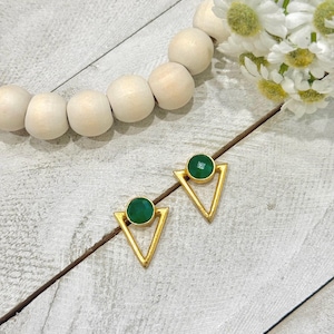 May include: Two gold-coloured triangle stud earrings, each with a round, emerald-green stone at the centre. The earrings are displayed on a light-coloured wooden surface, with a string of wooden beads and white flowers in the background.