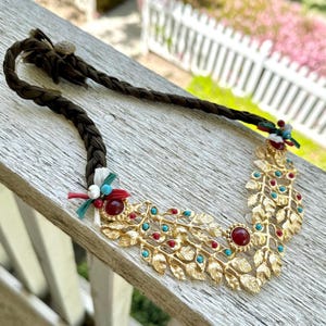 May include: A gold necklace with a leafy design and red and turquoise gemstones. The necklace is on a wooden surface.