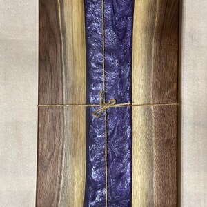 May include: A rectangular wooden chopping board with a purple resin inlay. The board is tied with twine and has a natural wood finish.