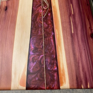 May include: A wooden cutting board with a purple and red resin inlay. The board is tied with twine and has a natural wood finish.