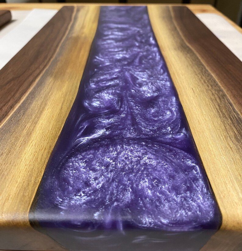 May include: A wooden chopping board with a purple and white resin inlay. The resin has a swirling pattern.