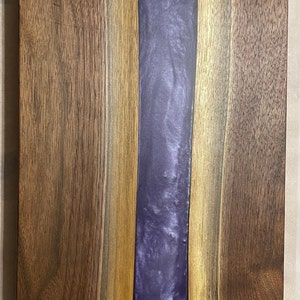 May include: A wooden chopping board with a purple resin inlay. The board is made of walnut wood and has a natural finish.