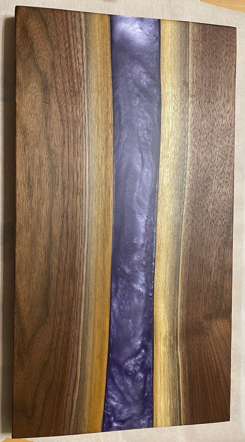 May include: A wooden chopping board with a purple resin inlay. The board is made of walnut wood and has a natural finish.