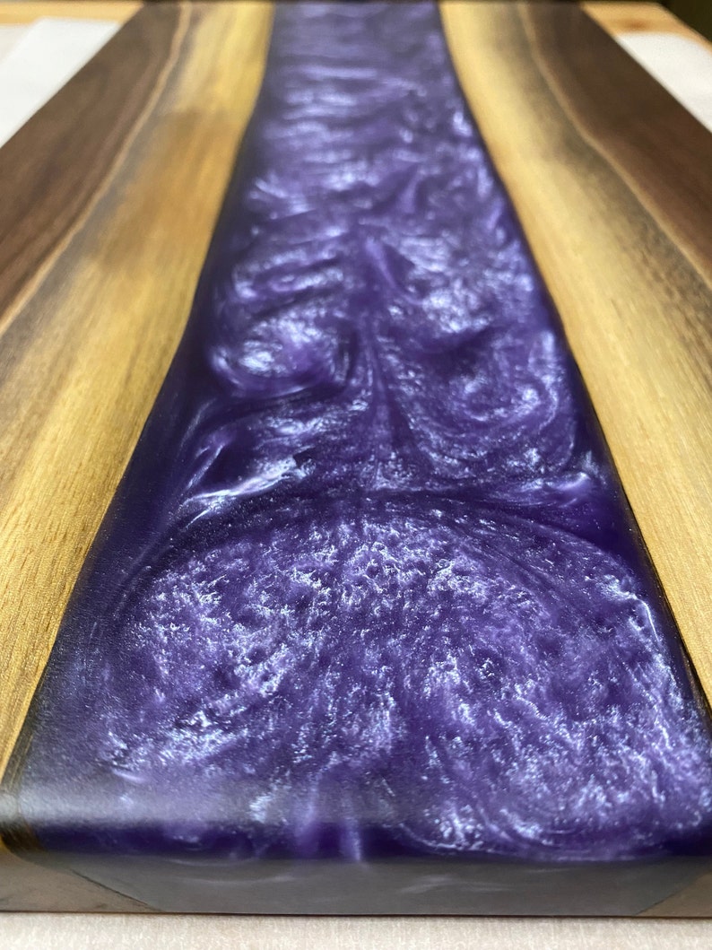 May include: A wooden chopping board with a purple resin inlay. The resin has a swirling pattern and a glittery finish.