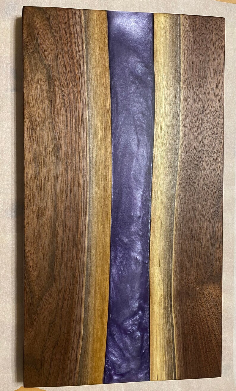 May include: A wooden chopping board with a purple resin inlay. The board has a natural wood finish and is made of walnut wood.