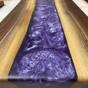 May include: A wooden chopping board with a purple and white epoxy resin inlay. The resin has a swirling pattern and a glossy finish.