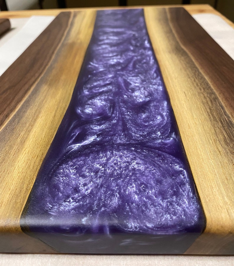 May include: A wooden chopping board with a purple and white epoxy resin inlay. The resin has a swirling pattern and a glossy finish.