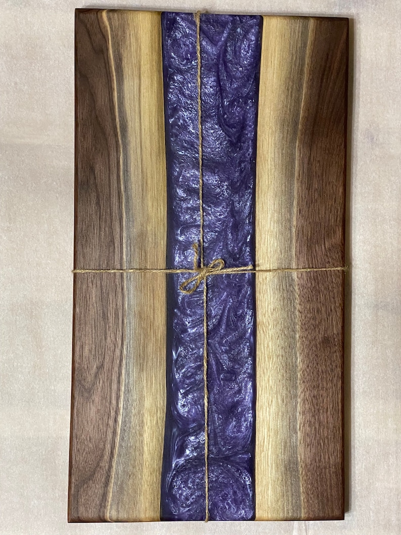 May include: A rectangular wooden chopping board with a purple resin inlay. The board is tied with twine and has a natural wood finish.