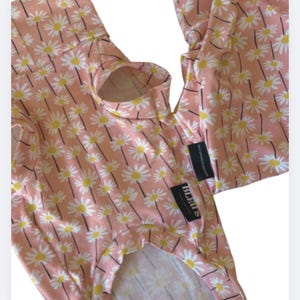 May include: A pink dog outfit featuring a daisy print. The garment has a turtleneck and openings for the front legs. The fabric is patterned with white daisies with yellow centres and black stems. The label reads "BLEUS".