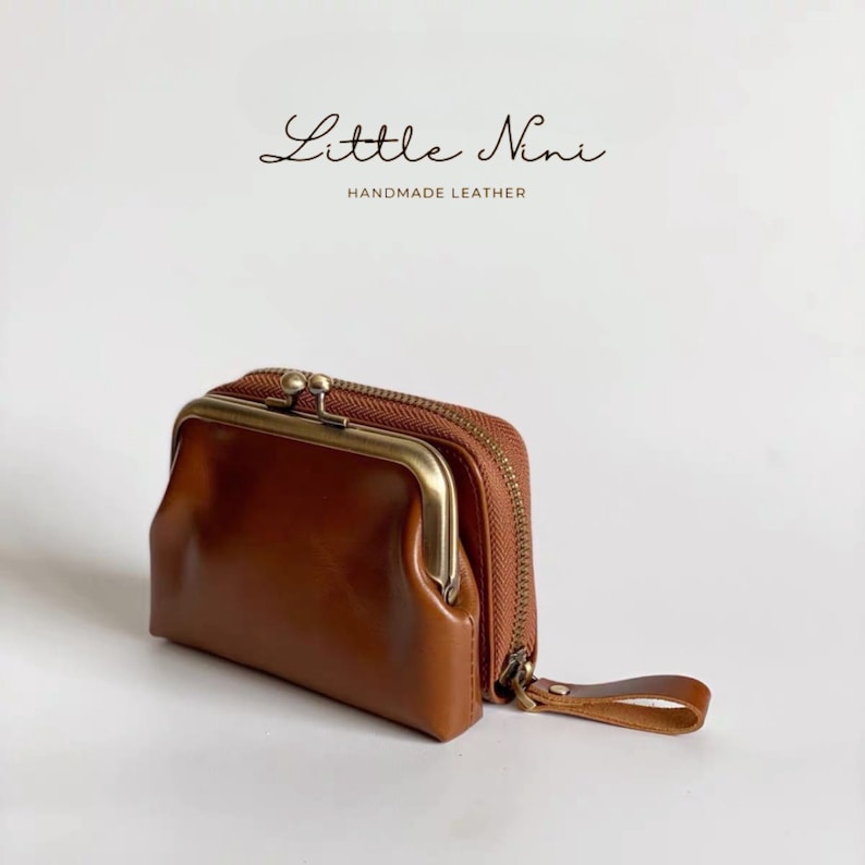 May include: A brown leather coin purse with a brass clasp and zipper closure. The purse has a small strap attached to the side. The purse is labeled "Little Nini Handmade Leather".