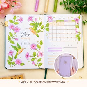 Op de afbeelding: Open journal met mei-kalender, kolibrie-illustraties en roze bloemaccenten. Het tijdschrift heeft een maandkalender, focus- en taaksecties en een kleine inzet van een lavendelkleurig tijdschrift. De tekst luidt "224 ORIGINELE HANDGETEKEND PAGINA'S."