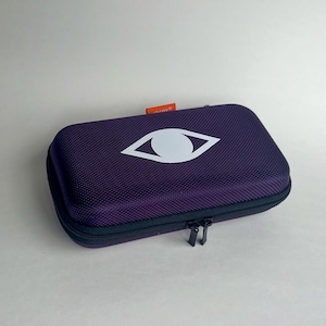 May include: A rectangular, purple case with a white eye logo. The case has a textured surface and a black zipper closure. A small orange tag is visible on the top. The case is designed to protect its contents.