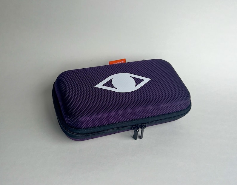 May include: A rectangular, purple case with a white eye logo. The case has a textured surface and a black zipper closure. A small orange tag is visible on the top. The case is designed to protect its contents.