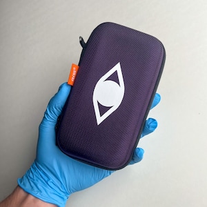 May include: A purple zippered case with a white eye-shaped logo. The case has a textured surface and an orange tag that reads "GLCON". The case is held in a blue gloved hand.