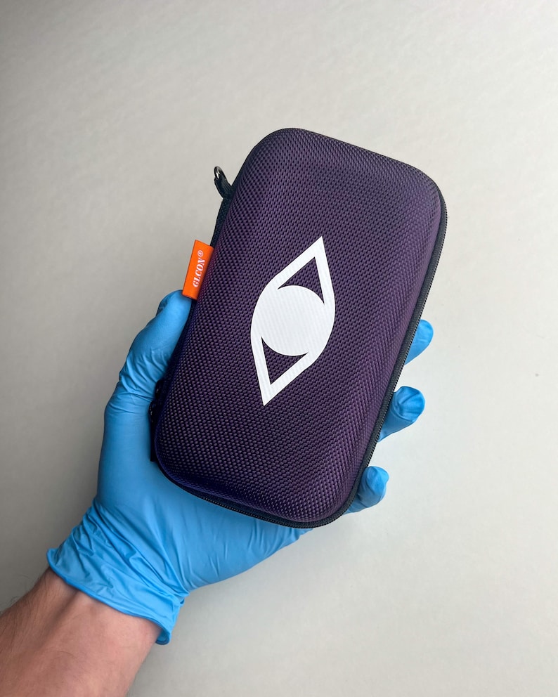 May include: A purple zippered case with a white eye-shaped logo. The case has a textured surface and an orange tag that reads "GLCON". The case is held in a blue gloved hand.