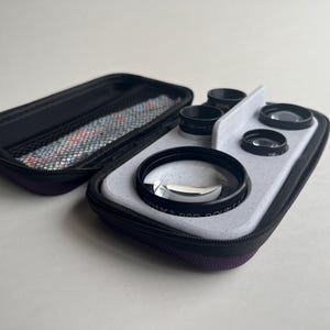 May include: A black and purple zippered case holds a set of camera lenses. The lenses are black with clear glass, and are arranged on a light gray tray. The case is open, revealing a patterned interior lining.