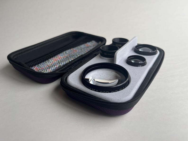 May include: A black and purple zippered case holds a set of camera lenses. The lenses are black with clear glass, and are arranged on a light gray tray. The case is open, revealing a patterned interior lining.