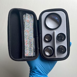 May include: A black zippered case, open to reveal a set of camera lenses and accessories. The case has a gray interior with various lens sizes and a mesh pocket containing a patterned cloth. The lenses are in a variety of sizes.