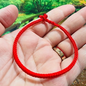May include: A red string bracelet with a knot at the top. The bracelet is tied around a person's wrist.
