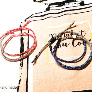 May include: Two braided bracelets, one with red, pink, and blue threads, and the other with blue, gray, and brown threads. The bracelets are on a fabric surface with the text "Do what you love".