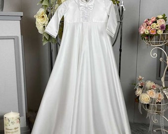 May include: White satin christening gown with long sleeves, a bow, and lace detailing. The gown is hanging on a hanger and has a white bonnet on top.