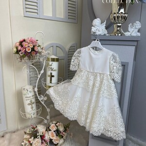 May include: White lace christening gown with a satin bodice and a tiered skirt. The gown is hanging on a white hanger and is displayed on a gray pedestal. The gown is decorated with delicate lace and has a scalloped hem.