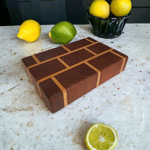 May include: A rectangular wooden cutting board with a brick-like pattern. The board is made of dark brown wood with lighter brown accents. Lemons and a lime are scattered around the board, with more lemons in a black bowl.