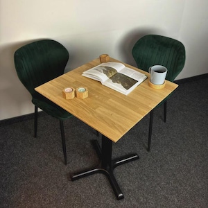 Könnte beinhalten: Ein quadratischer Tisch aus hellem Eichenholz mit schwarzem Säulenfuß, dazu zwei mit dunkelgrünem Samt bezogene Stühle. Auf dem Tisch liegen ein aufgeschlagenes Buch, eine hellgraue Keramiktasse auf einem Holzuntersetzer und zwei kleine Holzkerzenhalter.