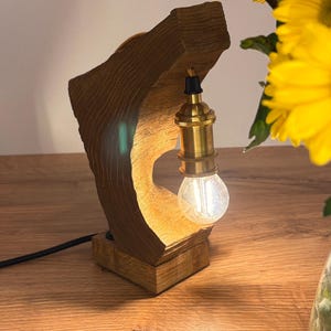 Può includere: Una lampada da tavolo in legno con una finitura in legno naturale e una lampadina rotonda in vetro trasparente. La lampada è su una base in legno e ha un cavo nero.