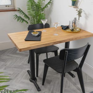 Puede incluir: Una mesa de comedor rectangular de madera clara con un tablero grueso y una base central negra, acompañada de dos sillas de madera negra. Sobre la mesa hay dos copas de martini transparentes, un mantel individual negro y un cuenco blanco.