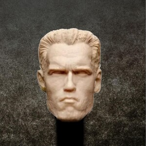 May include: A close-up of a light brown unpainted plastic head sculpt of a man with short, wavy hair. The head sculpt has a serious expression with furrowed brows and a slightly open mouth.