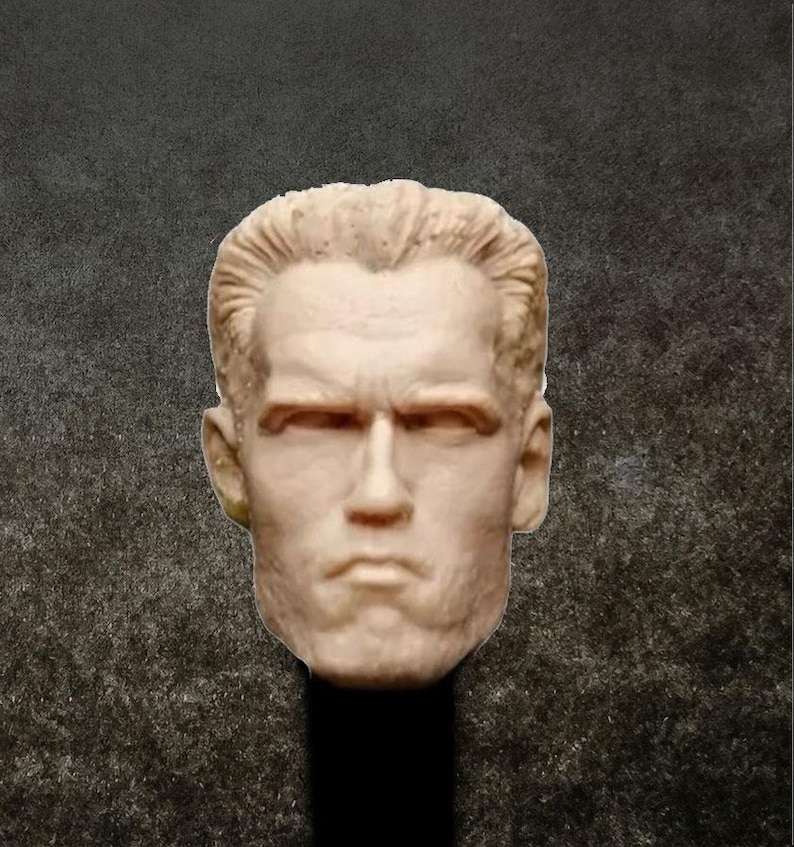 May include: A close-up of a light brown unpainted plastic head sculpt of a man with short, wavy hair. The head sculpt has a serious expression with furrowed brows and a slightly open mouth.