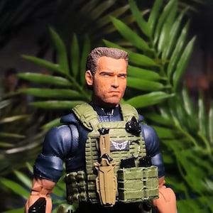 May include: A military action figure wearing a green tactical vest, a black knife, and a black pistol. The figure has short brown hair and a serious expression.