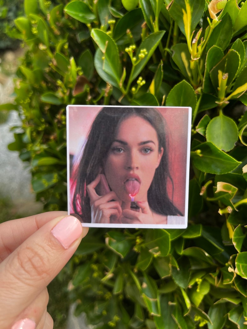 May include: A close-up of a woman's face with her tongue sticking out and a lighter in her hand. The background is blurred and pink.