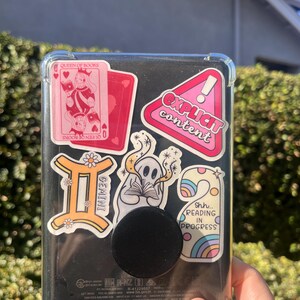 May include: A black e-reader with a clear protective case, adorned with colorful stickers. Stickers include a pink playing card with "Queen of Books", a Gemini symbol, and a pink sticker that says "Explicit Content". A black pop socket is attached.