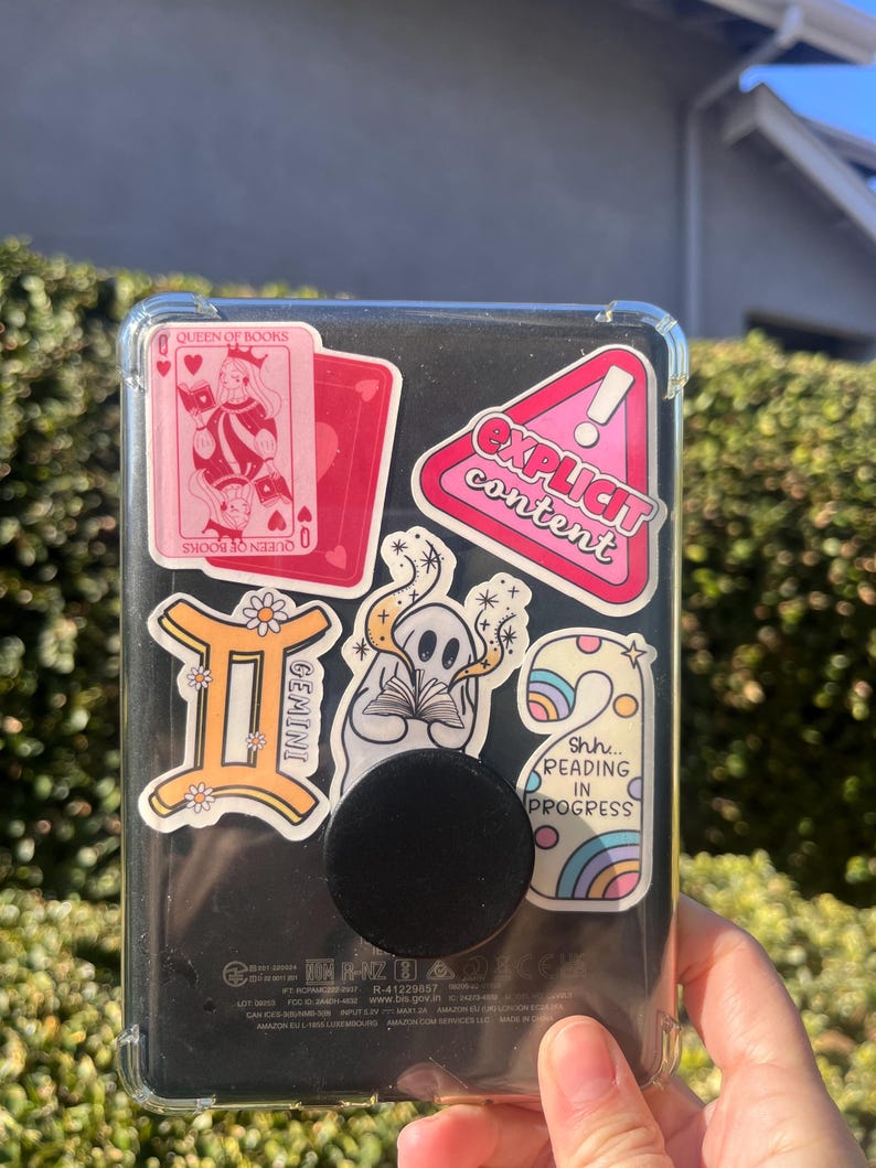 May include: A black e-reader with a clear protective case, adorned with colorful stickers. Stickers include a pink playing card with "Queen of Books", a Gemini symbol, and a pink sticker that says "Explicit Content". A black pop socket is attached.