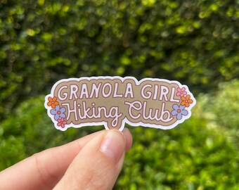 May include: A brown sticker with the text "Granola Girl Hiking Club" in white cursive lettering. The sticker is surrounded by small pink, blue, and orange flowers.