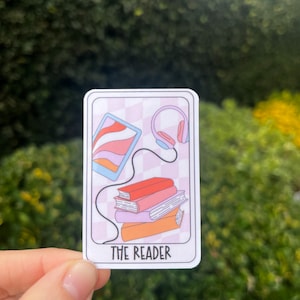 May include: A sticker with a pink, white, and blue illustration of a tablet, headphones, and a stack of books. The text "THE READER" is printed at the bottom of the sticker.
